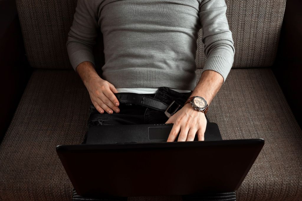 A man is sitting on a sofa with a belt and trousers zipper unfastened and is keeping the laptop on his knees.