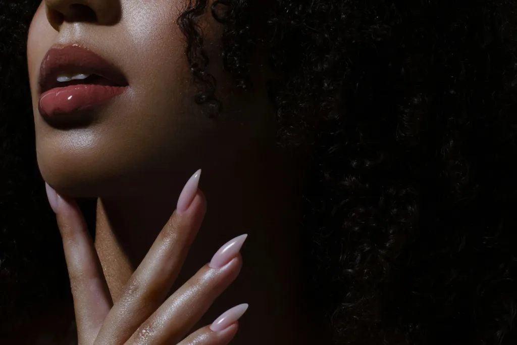A woman with sparkling lips and a slightly opened mouth but with a hidden face is gently touching with fingers in pink manicure her chin.