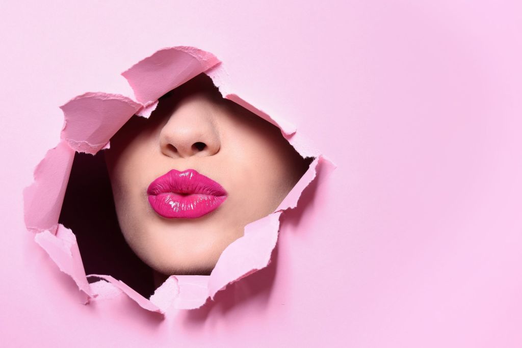 A woman with only visible lips and pink lipstick sends air kisses through the hole-turned-pink cardboard.