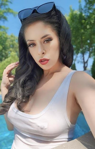 The Atomic Goddess is outdoors with black sunglasses holding her hair and a white semi-transparent top with visible nipples.
