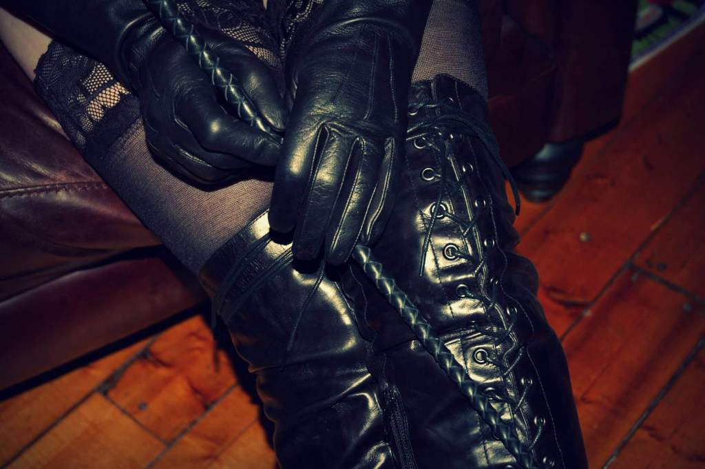 Mistress in black leather gloves and black leather high-heel boots holding a black leather whip.