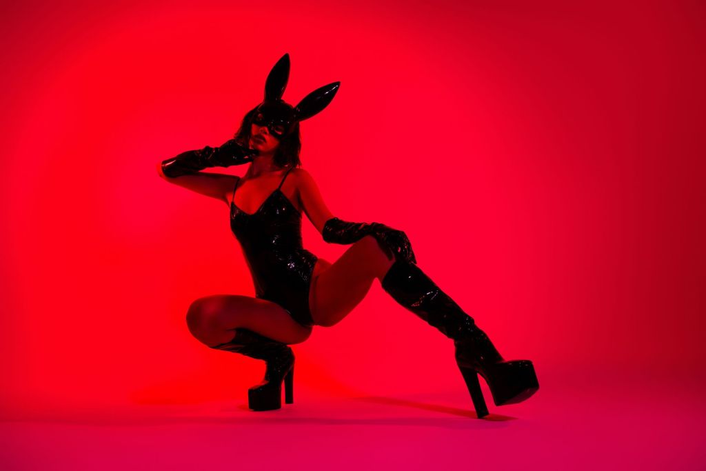 A female creator in a black latex bodysuit, black latex rabbit mask, knee-high black boots and elbow-high gloves is taking her shoots with fetish content