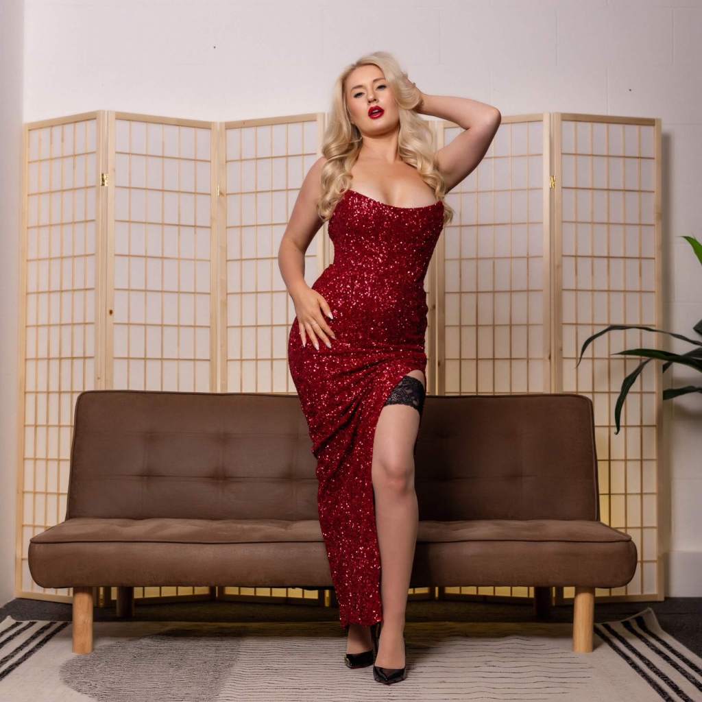 Miss Mae poses in a long red evening dress and exposes one leg in black stockings from the dress's cut